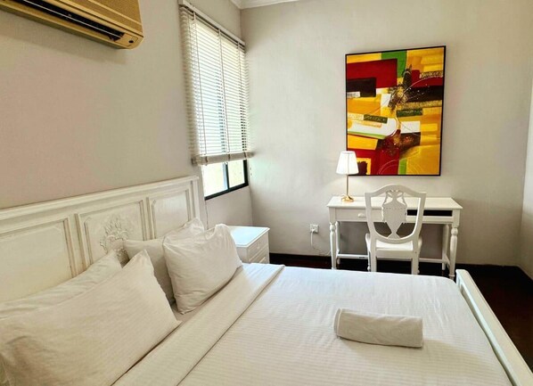 A hotel room with a bed, white bedding, a desk with a chair, a lamp, and a colorful abstract painting on the wall. - Century Bay Private Residences (George Town)