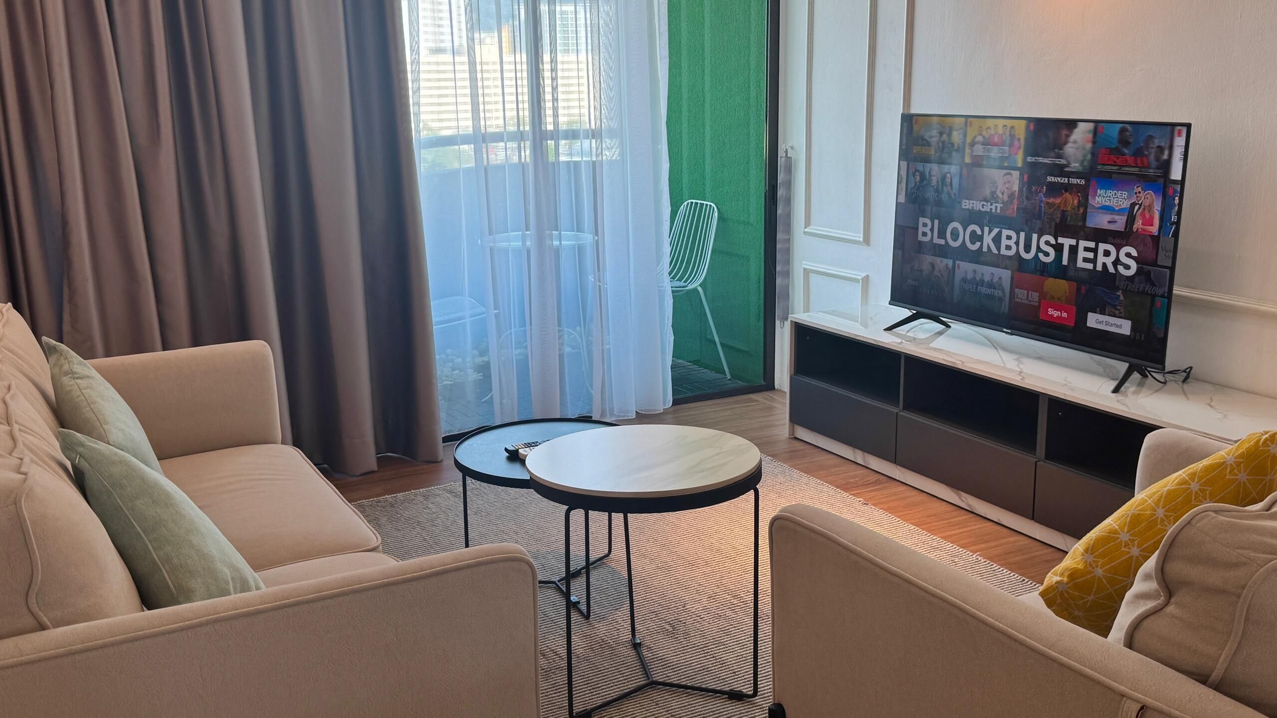 Ett modernt vardagsrum med en beige soffa, ett litet runt bord och en tv-skärm som visar programmet ”Blockbusters”.