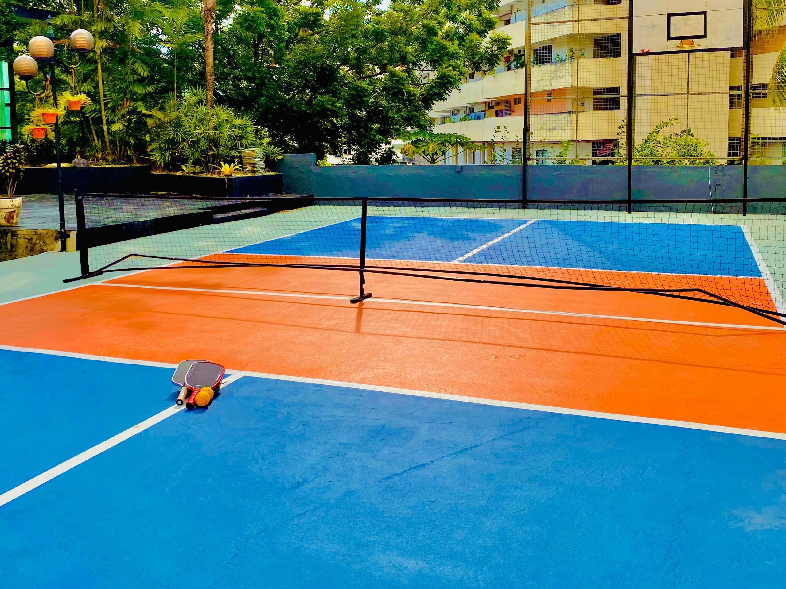 Un court de tennis en plein air, doté d’un filet, entouré de verdure et avec un bâtiment en arrière-plan.
