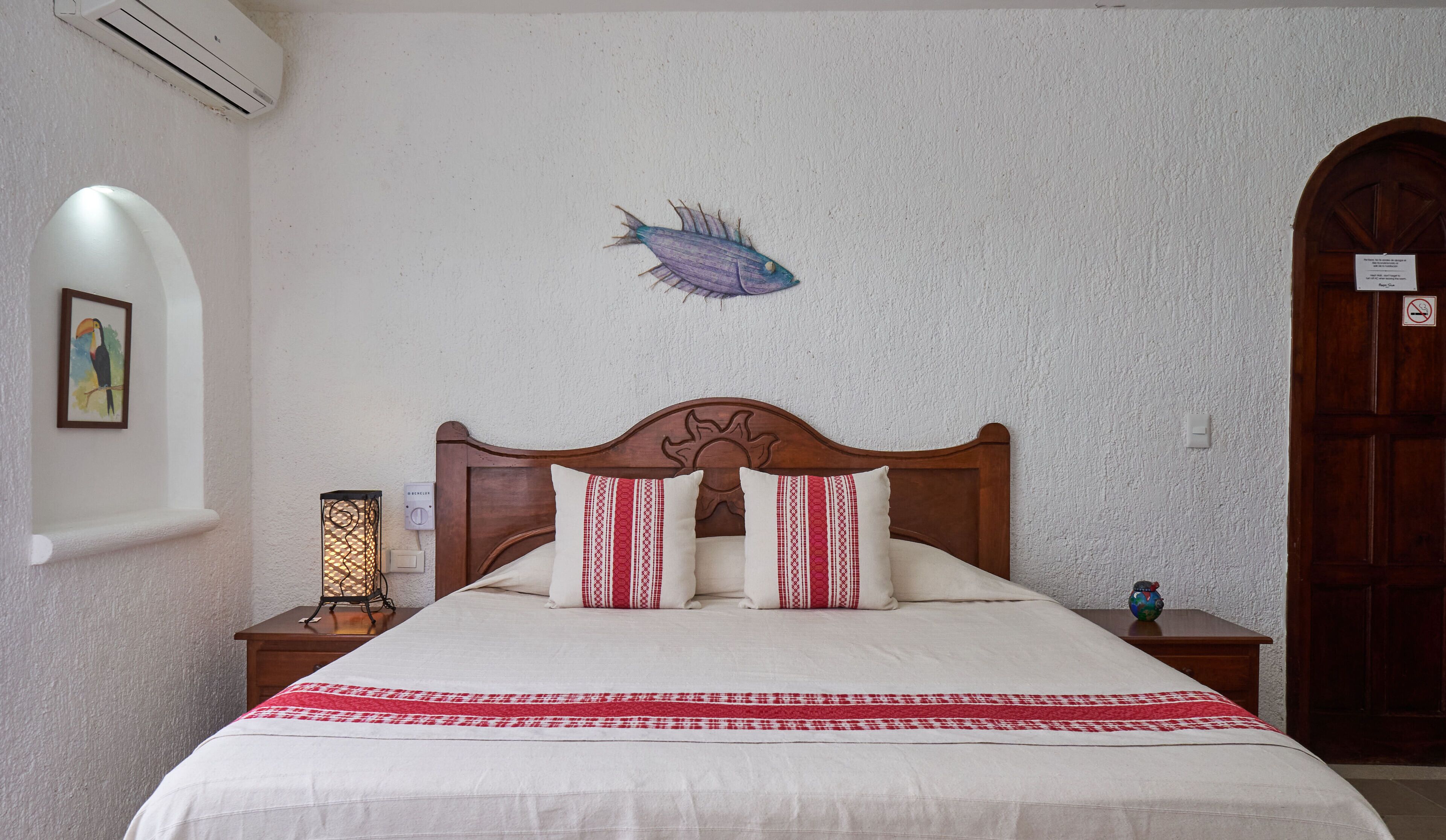 A bedroom with a wooden headboard, a bed with white and red bedding, a wall-mounted air conditioner, a wall-mounted lamp, and a framed picture.