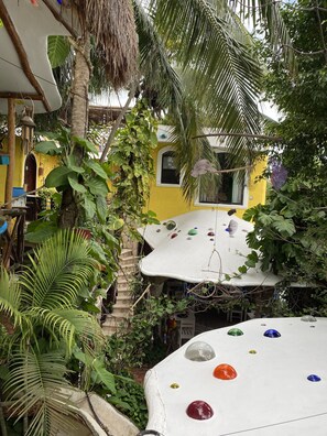 A tropical garden with a white table covered in colorful glass marbles, surrounded by lush greenery and a yellow building with a window.