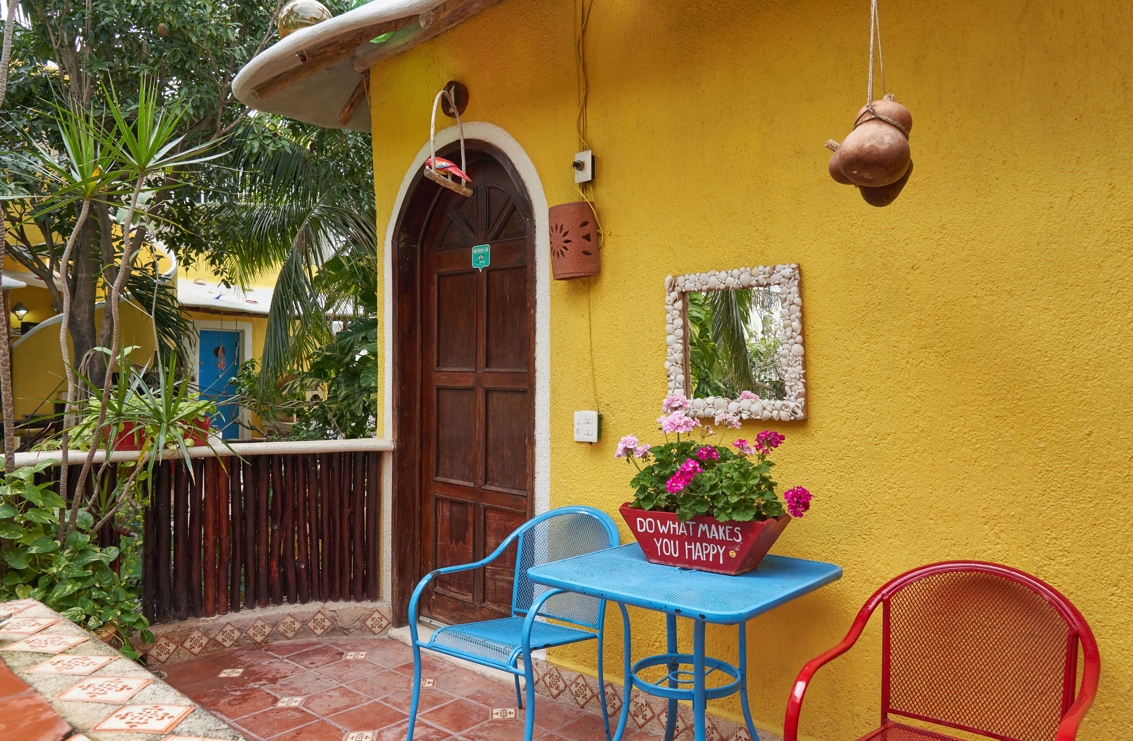 Un edificio amarillo con una puerta de madera, una mesa azul con una silla roja y una maceta con un cartel.