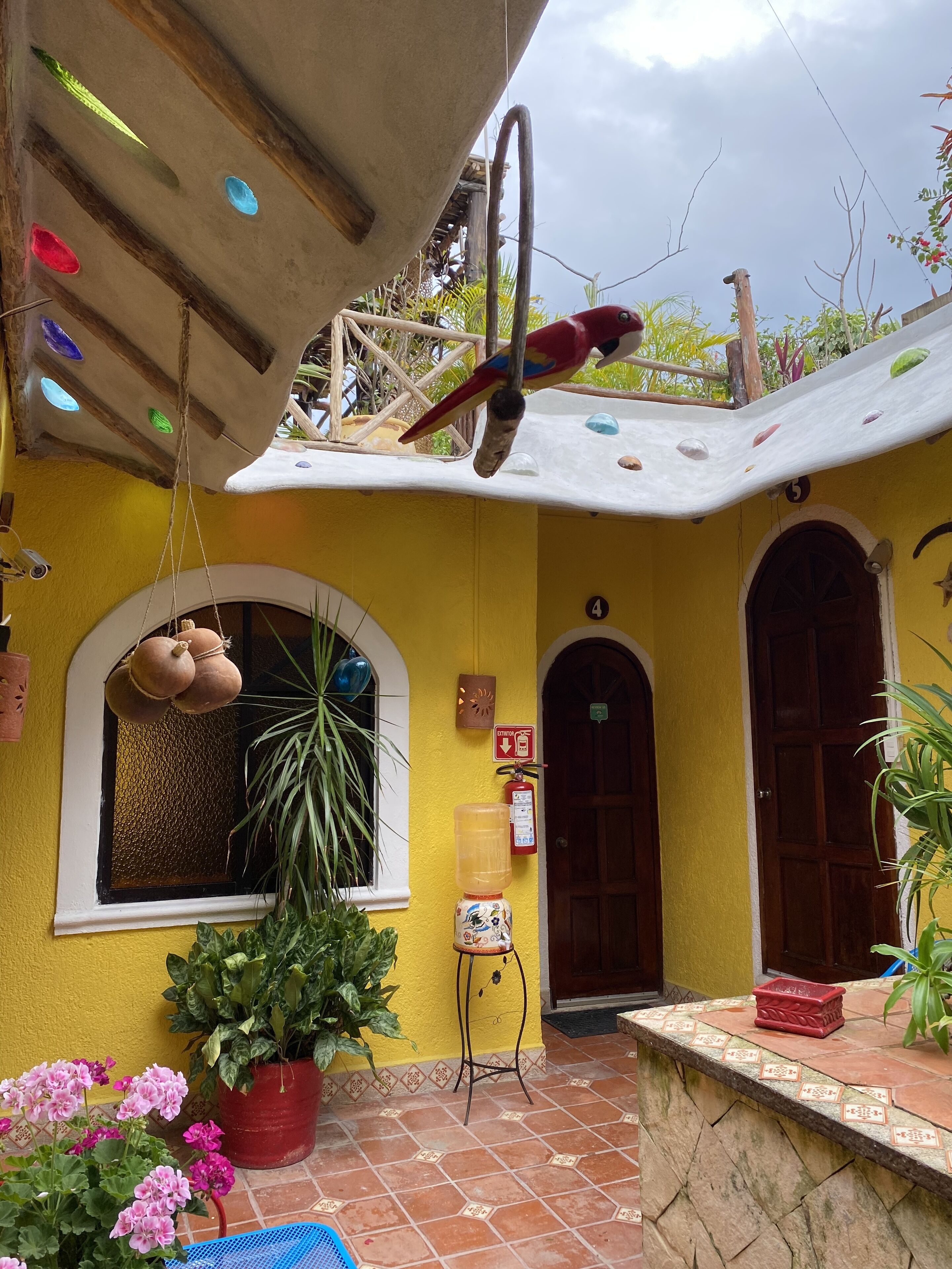 A yellow building with a tiled patio, a hanging birdcage, a potted plant, and a fire extinguisher.