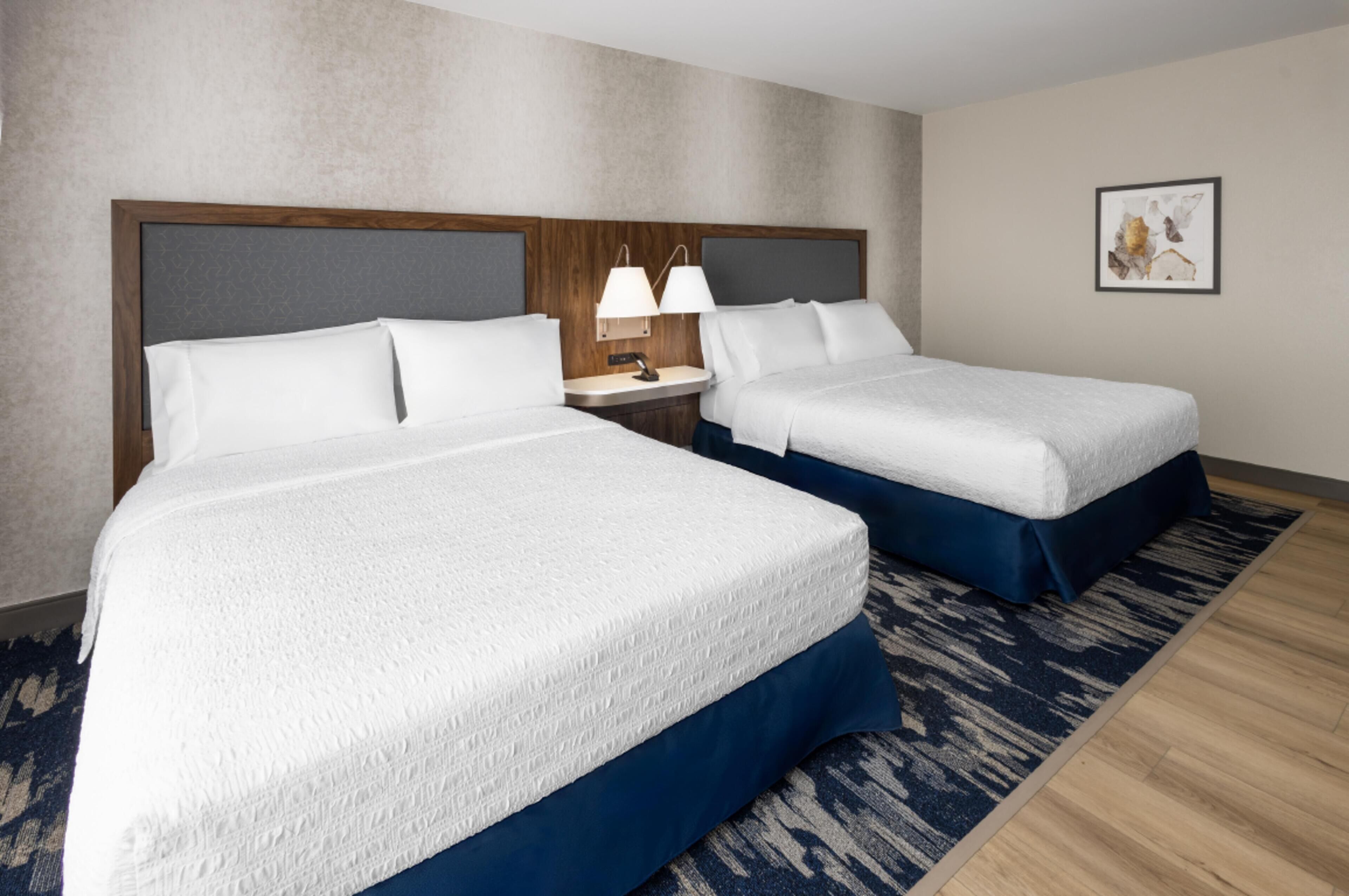 A hotel room with two beds, a nightstand with lamps, a framed picture on the wall, and a patterned rug.
