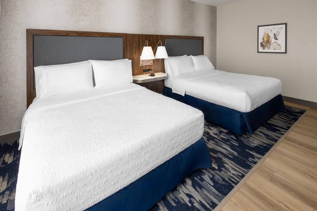 A hotel room with two beds, a wooden headboard, a nightstand with lamps, a patterned rug, and a framed picture on the wall.