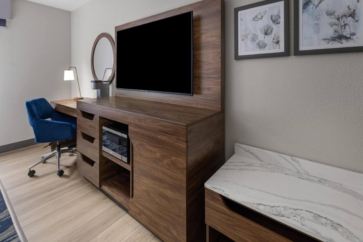 A modern hotel room with a wooden entertainment unit, a flat-screen TV, a blue chair, a bedside table, and two framed artworks on the wall.