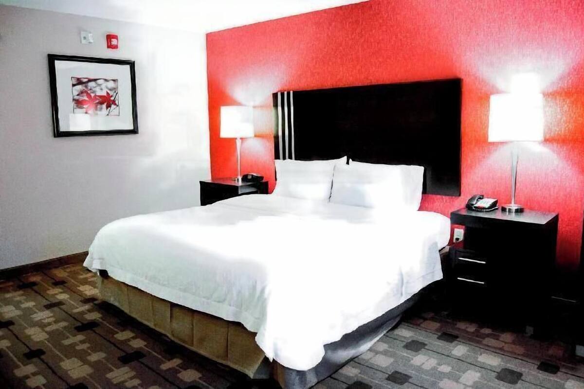 A hotel room with a bed, two bedside tables, a red accent wall, a framed picture, and a patterned floor.