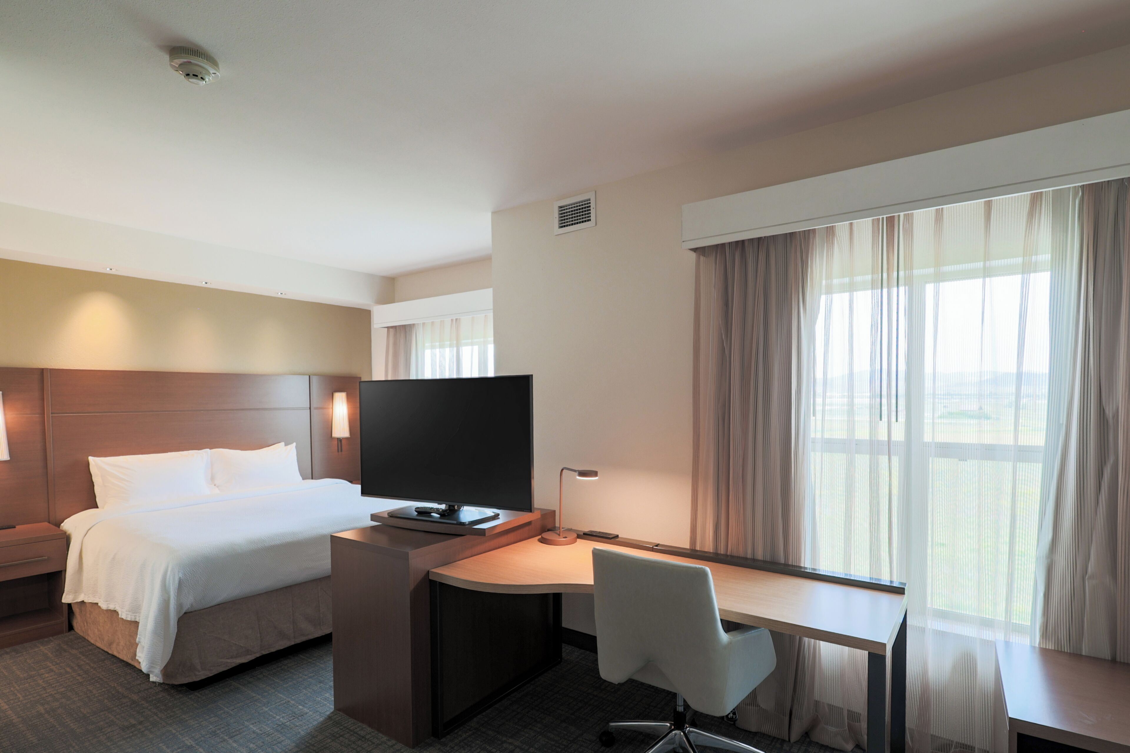 A hotel room with a bed, a desk with a chair, a television, and a large window with curtains.