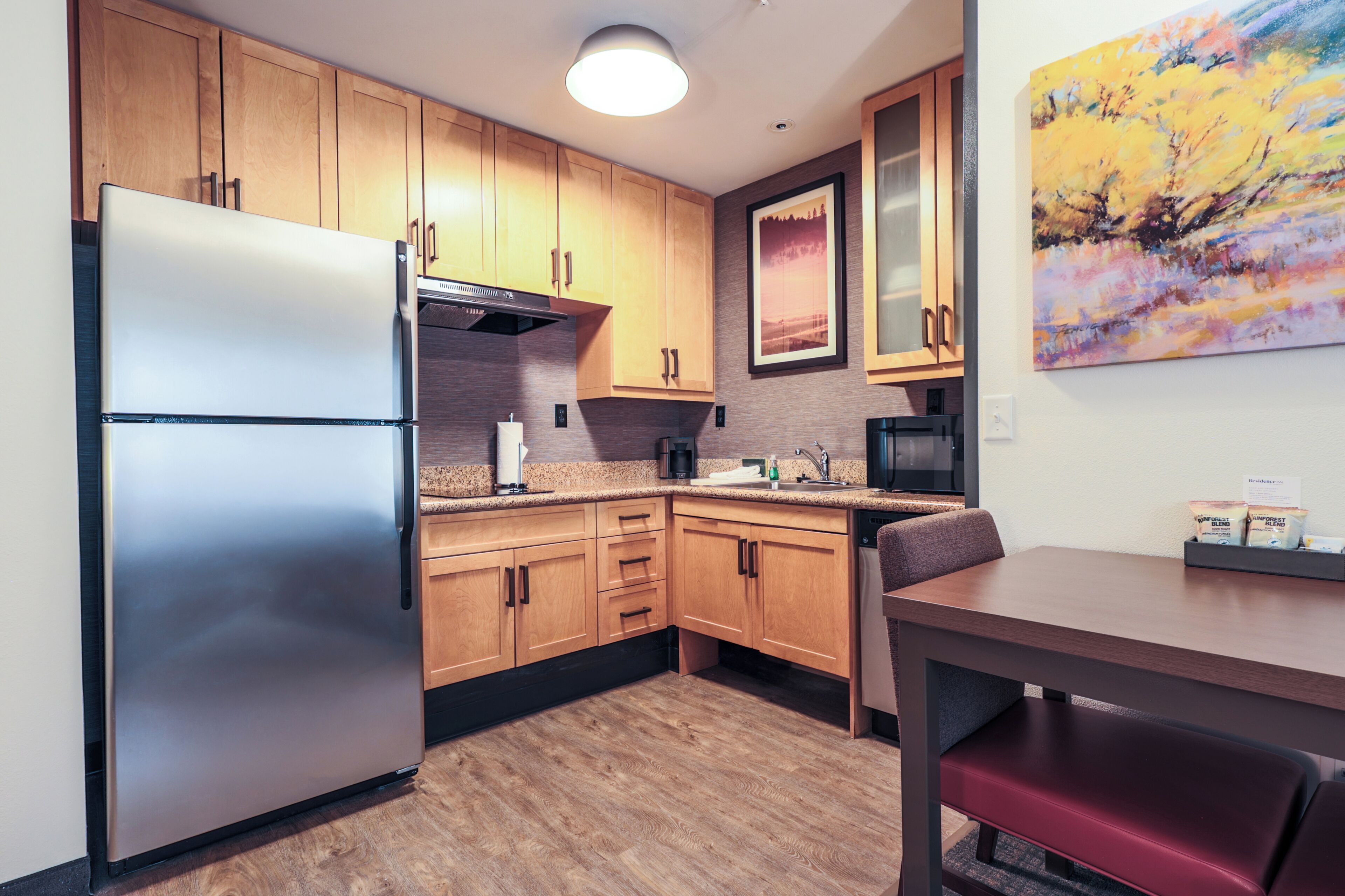 A compact kitchen with wooden cabinets, a stainless steel refrigerator, a microwave, and a dining area with a table and bench.