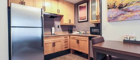 A compact kitchen with wooden cabinets, a stainless steel refrigerator, a microwave, and a dining area with a table and bench.