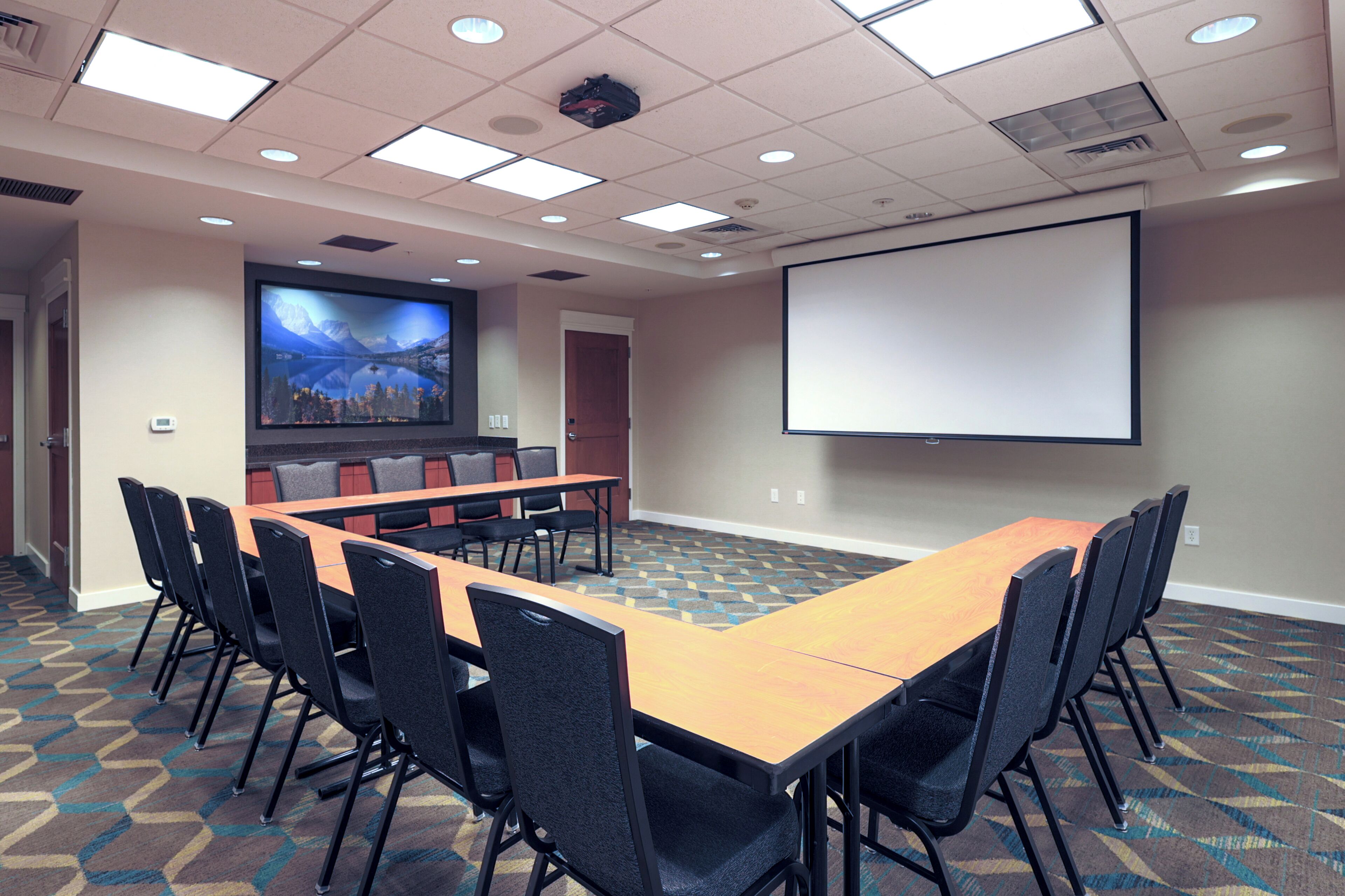 A conference room with a long table, chairs, a projector screen, and a large screen displaying a scenic image.