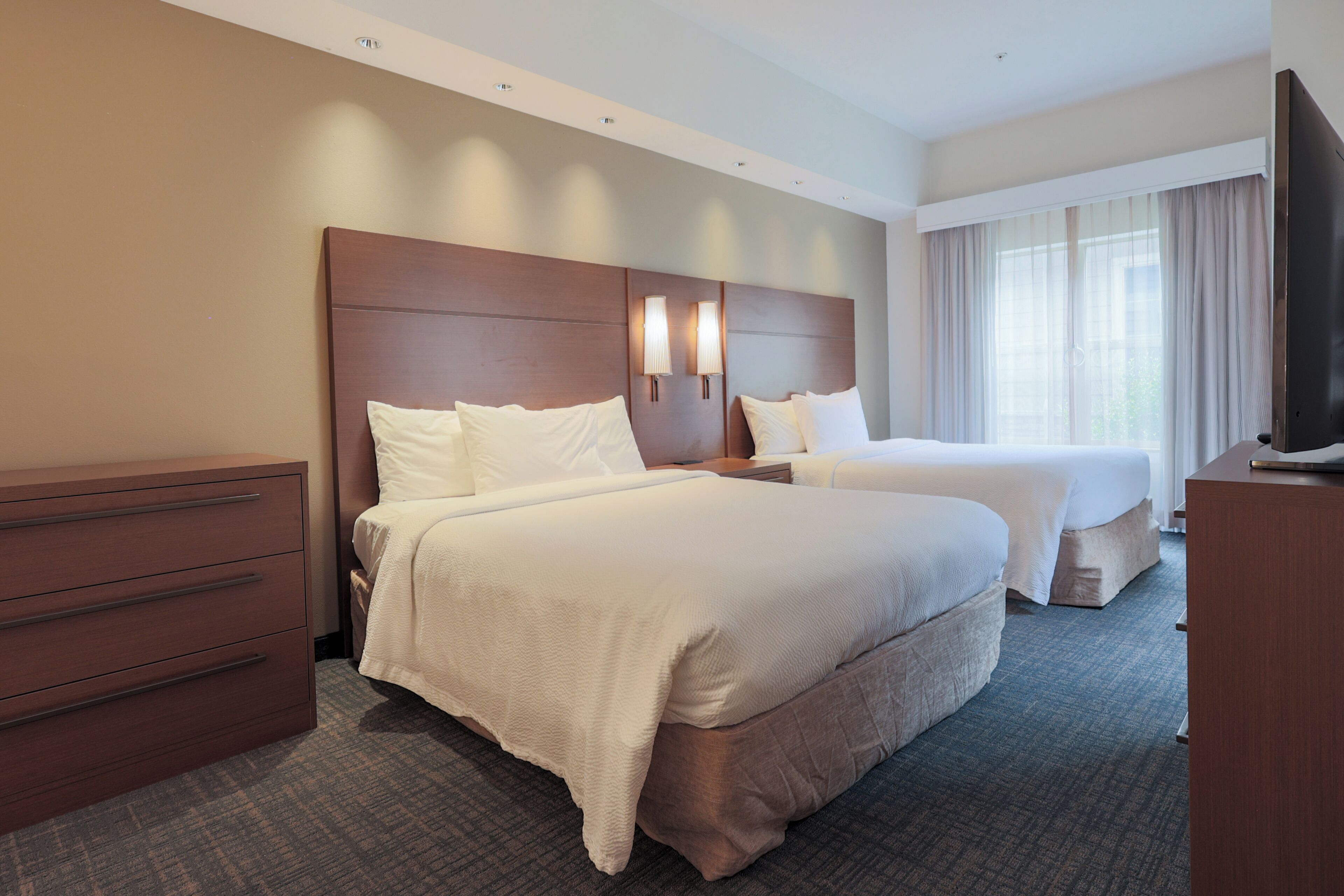 A hotel room with two beds, a dresser, a TV, and a window with curtains.