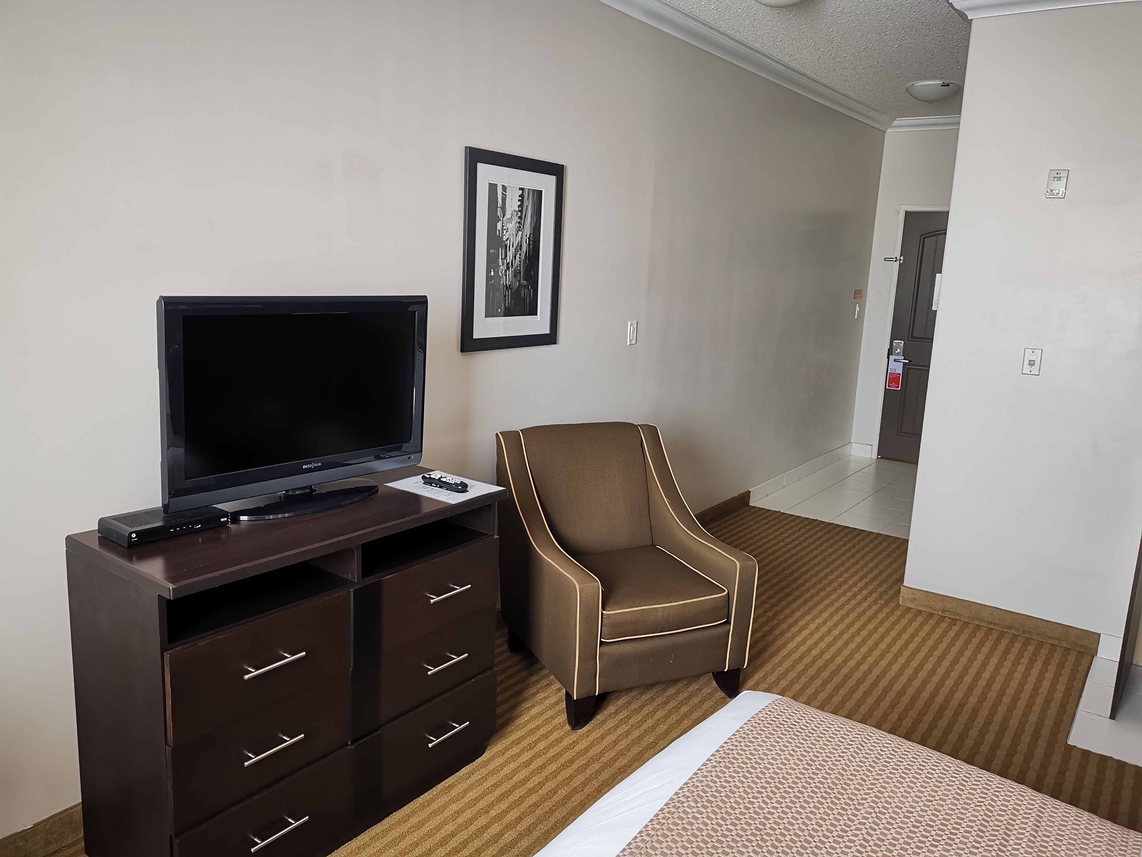 A hotel room with a dark wooden dresser, a flat-screen TV, a brown leather chair, a framed picture on the wall, and a carpeted floor.