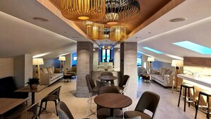 A modern hotel lobby with a central seating area, multiple tables, and a high ceiling with unique lighting fixtures. - Aparthotel Acuazul (Peniscola)