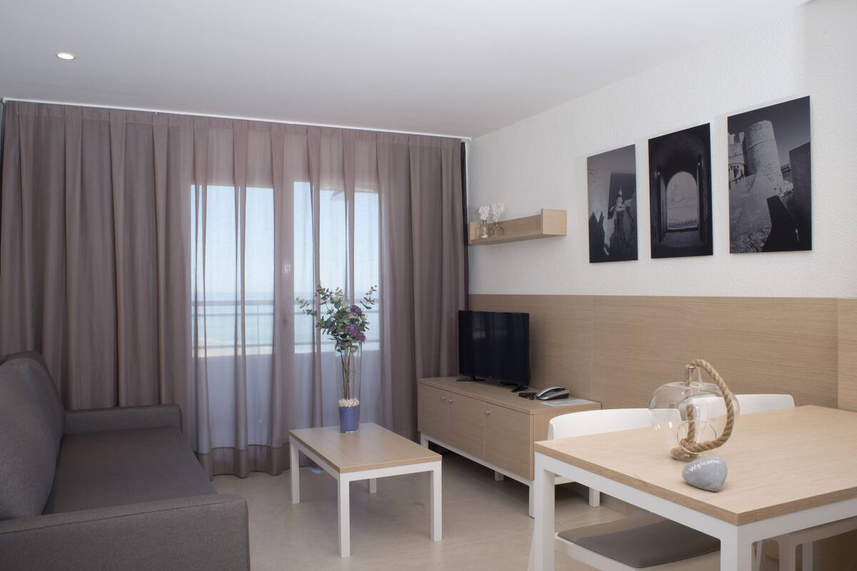Una habitación de hotel moderna con sofá, una mesa de comedor, televisión y un amplio ventanal con cortinas.