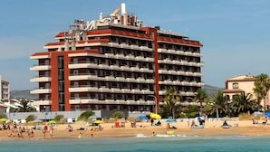 Un hotel de varios pisos con balcones, cerca de una playa con sombrillas y gente.