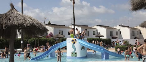 Zona de piscina con tobogán de agua, palmeras y edificios al fondo.