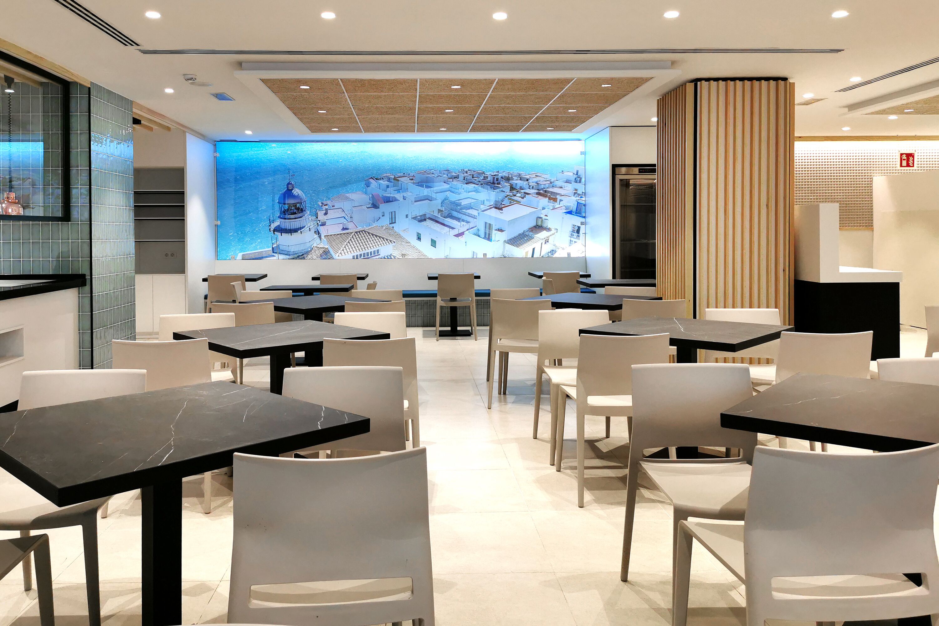 Un restaurante moderno con una gran pantalla digital mostrando un paisaje urbano, sillas y mesas blancas, y un diseño interior limpio y contemporáneo.