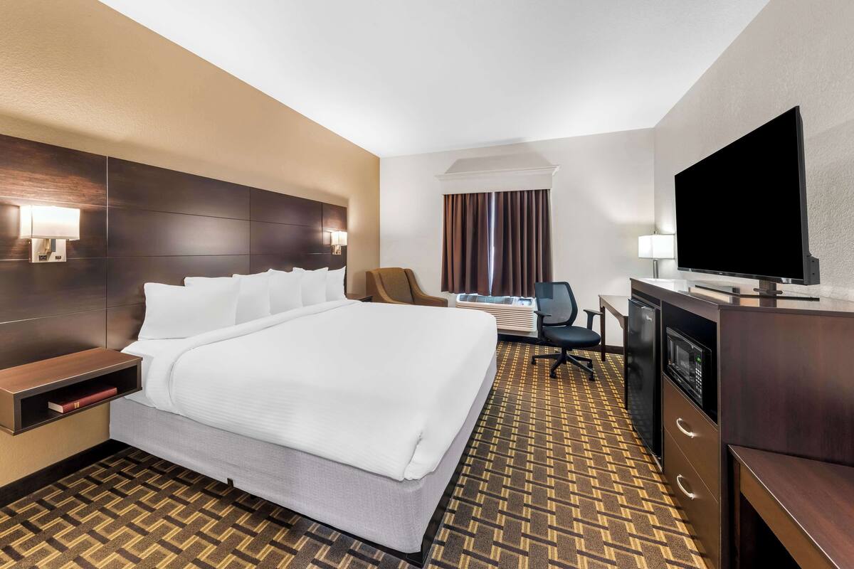 A hotel room with a large bed, a desk with a flat-screen TV, a chair, a desk lamp, and a minibar.