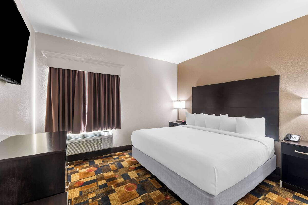 A hotel room with a large bed, a flat-screen TV mounted on the wall, a window with curtains, a bedside table with a lamp, and a patterned carpet.