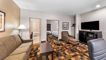 Una habitación de hotel con sofá, sillón, reposapiés, mesa de centro, televisión de pantalla plana y microondas.