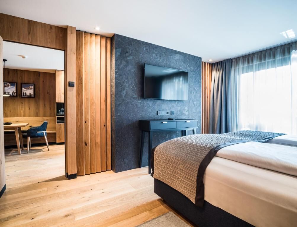 Une chambre d’hôtel moderne avec un plancher en bois, un lit recouvert d’une courtepointe à motifs, un téléviseur à écran plat fixé à un mur foncé et un bureau avec une chaise.