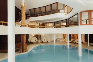 Un área de piscina interior con varios niveles, piscina curvada, columnas de madera y sillones de descanso.