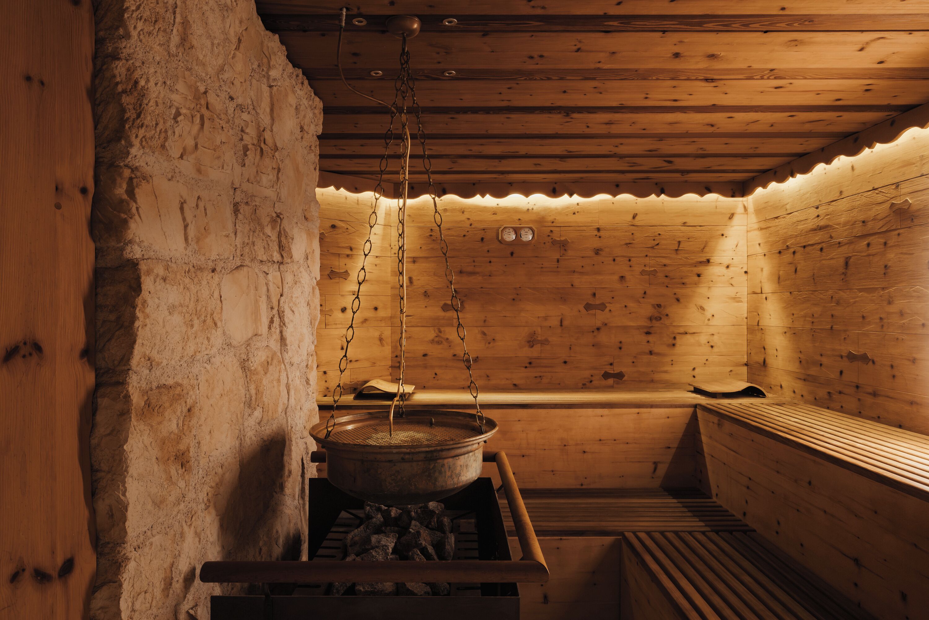 A sauna with wooden walls and benches, a stone wall on one side, and a large pot on a stove.