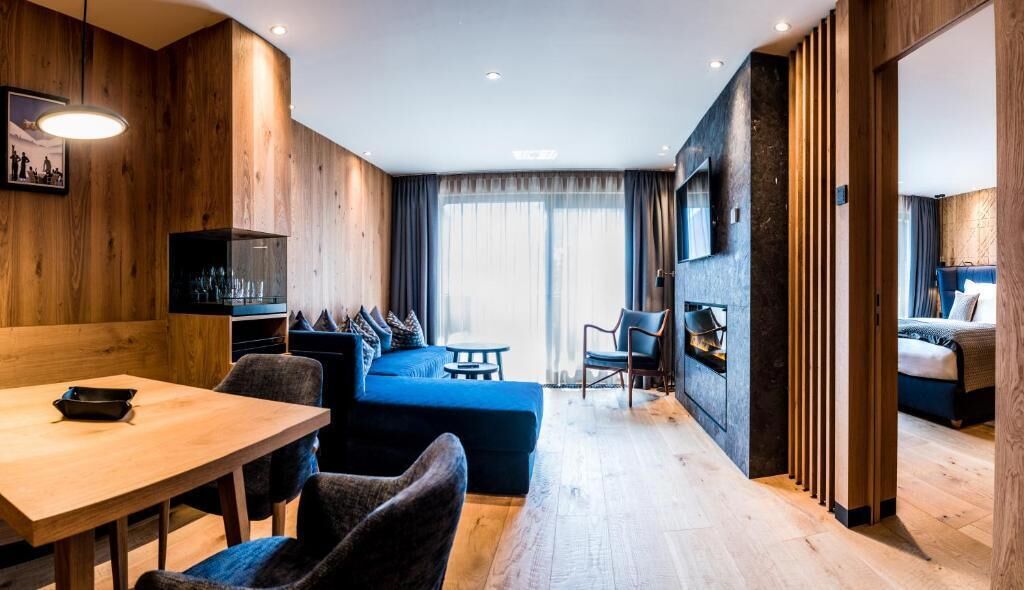 A modern hotel room with a wooden accent wall, a dining area with a table and chairs, a blue sofa, and a separate sleeping area with a bed.