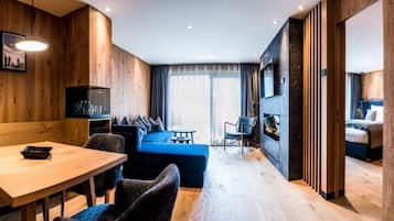 A modern hotel room with a wooden accent wall, a dining area with a table and chairs, a blue sofa, and a separate sleeping area with a bed.