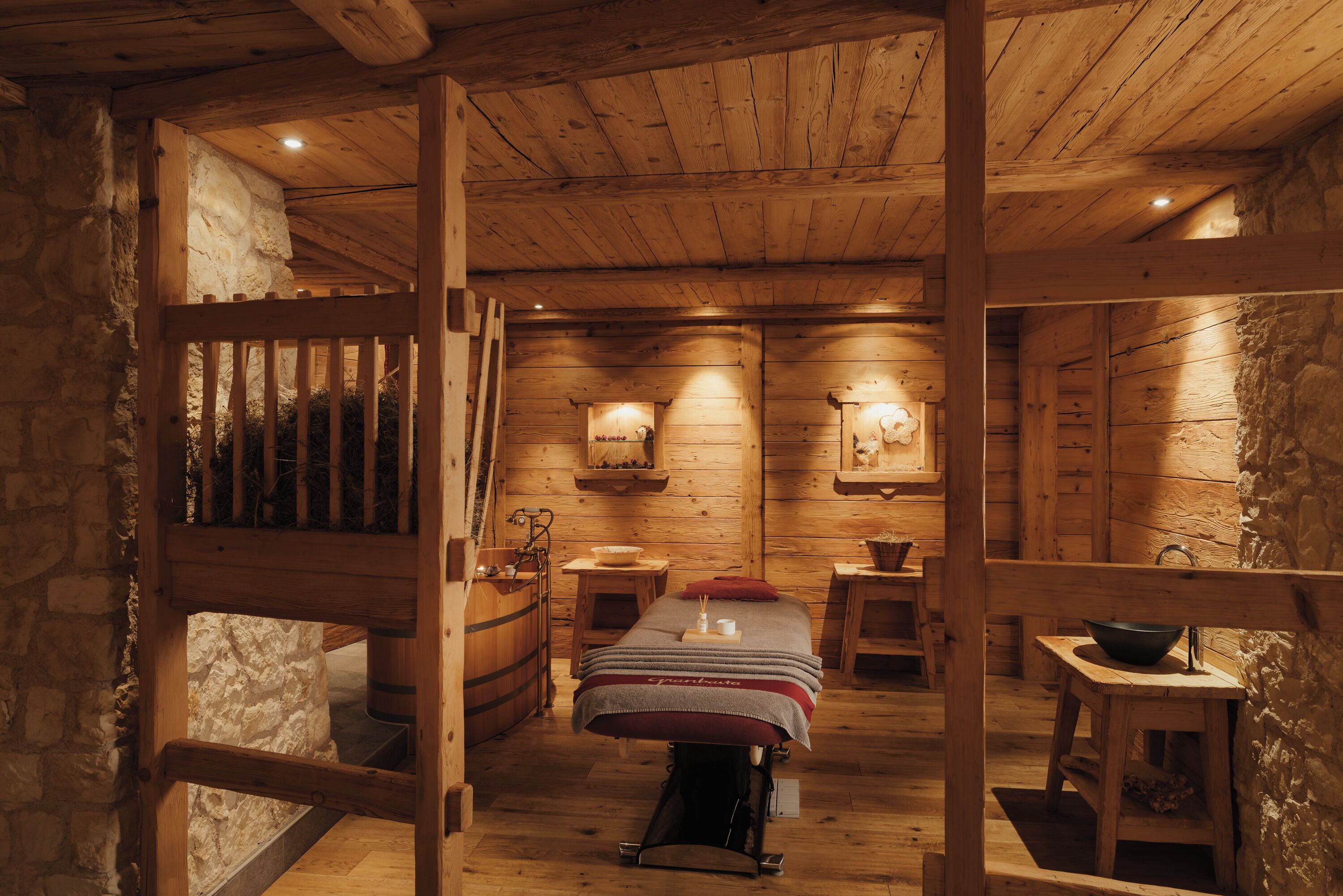 Interior de una cabaña de madera con una cama central, paredes de piedra y muebles rústicos.