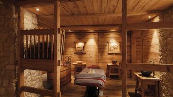 Interior de una cabaña de madera con una cama central, paredes de piedra y muebles rústicos.