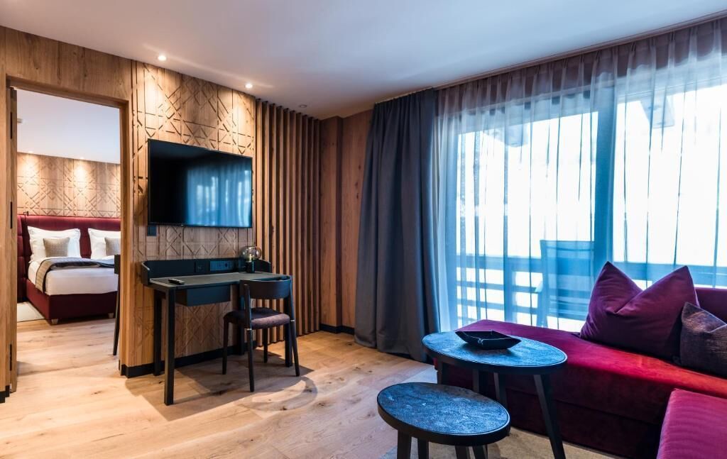 Habitación de hotel con cama, escritorio y silla, una mesita, sofá y un ventanal con cortinas.