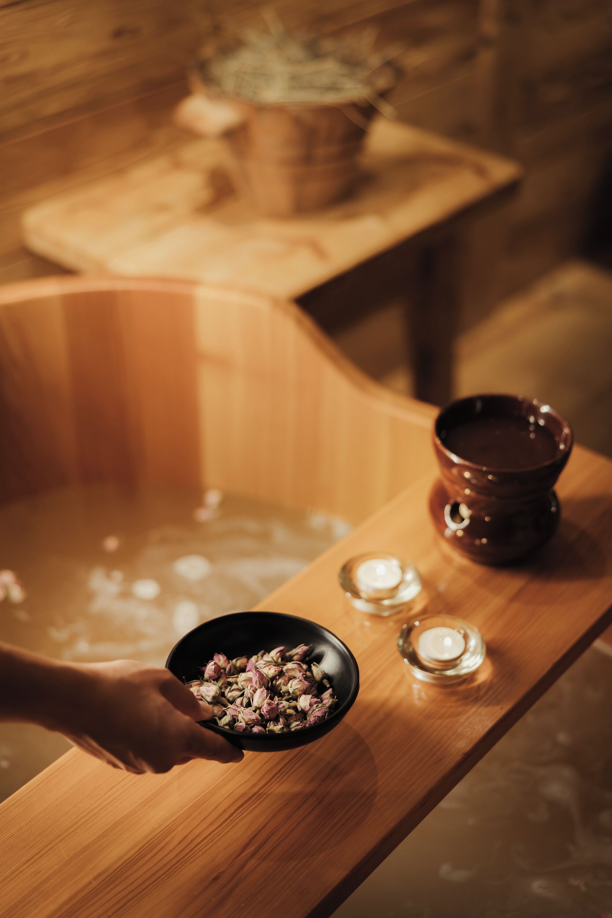 Une baignoire en bois dans laquelle une personne tient un bol rempli de fleurs. On peut voir des bougies et un petit bol sur une table en bois, à côté de la baignoire.