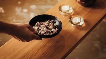 Une baignoire en bois dans laquelle une personne tient un bol rempli de fleurs. On peut voir des bougies et un petit bol sur une table en bois, à côté de la baignoire.