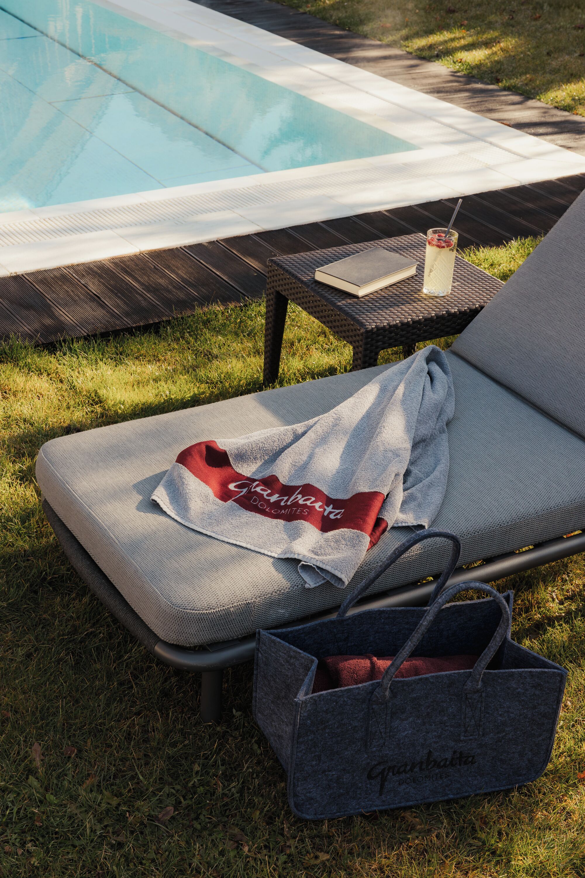 A poolside lounge chair with a towel and a bag, a glass of drink, and a book on a table.