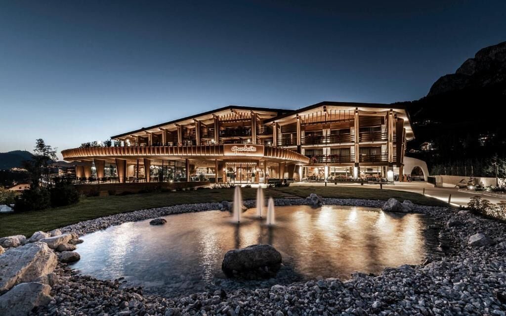 A multi-story hotel with a modern architectural design, featuring a large outdoor pool with a fountain, surrounded by a rocky landscape.