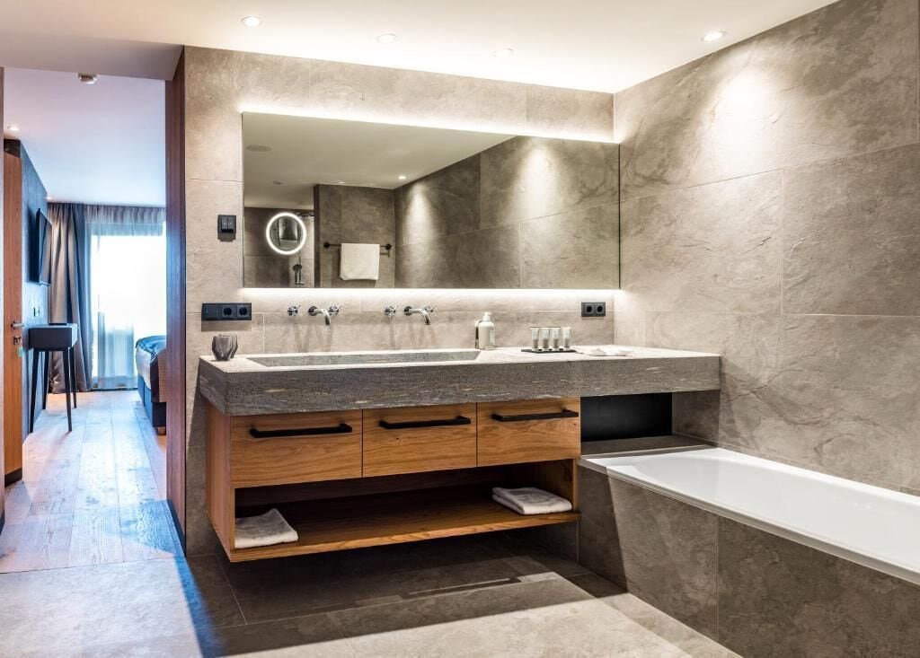 A modern bathroom with a large mirror, two sinks, a bathtub, and a wooden vanity.