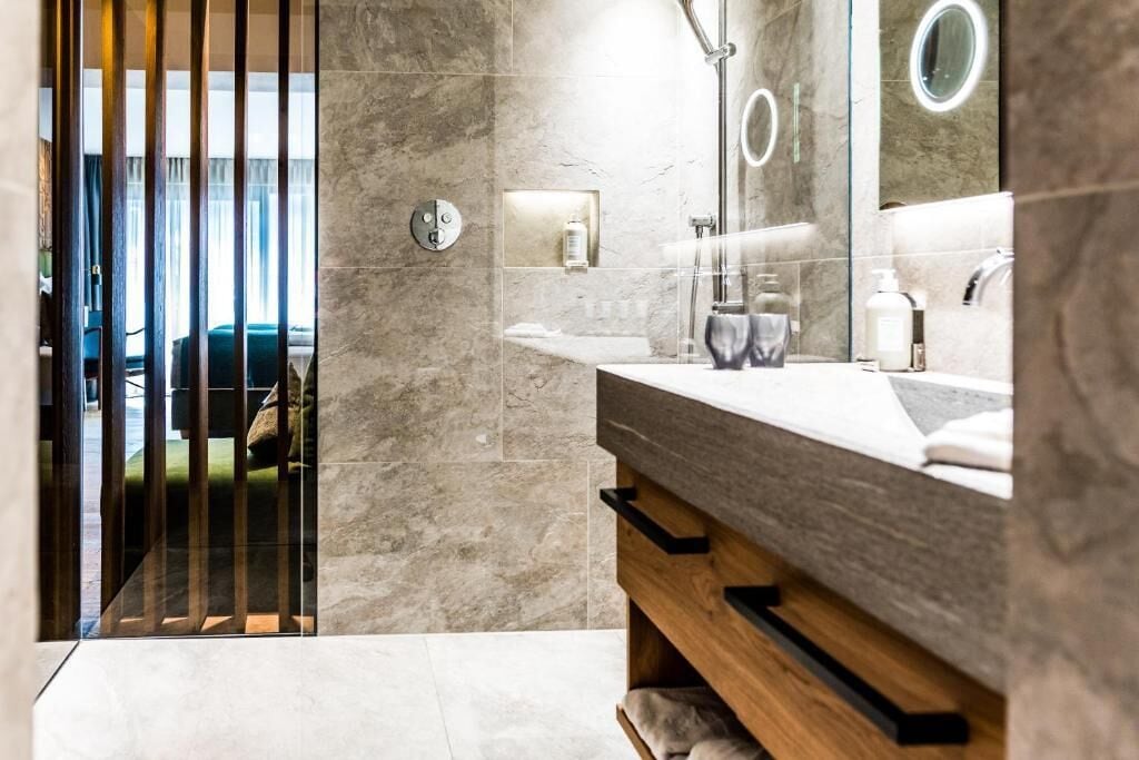 Een moderne badkamer met een marmeren vloer, een houten wastafelmeubel, een grote spiegel met ronde lijst en een douchegedeelte.