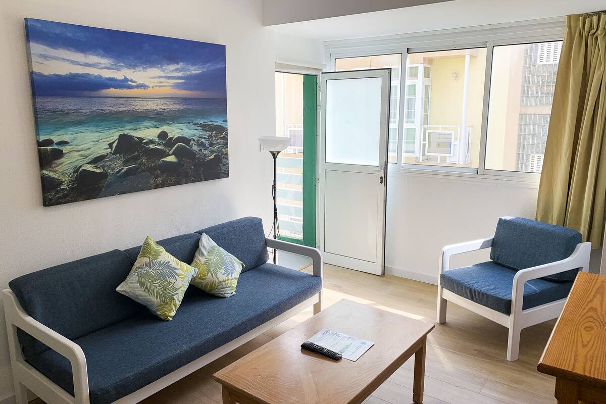 A room with a blue sofa, a wooden coffee table, and a white armchair. There is a painting of a beach scene on the wall, and a sliding glass door leading to an outdoor area.