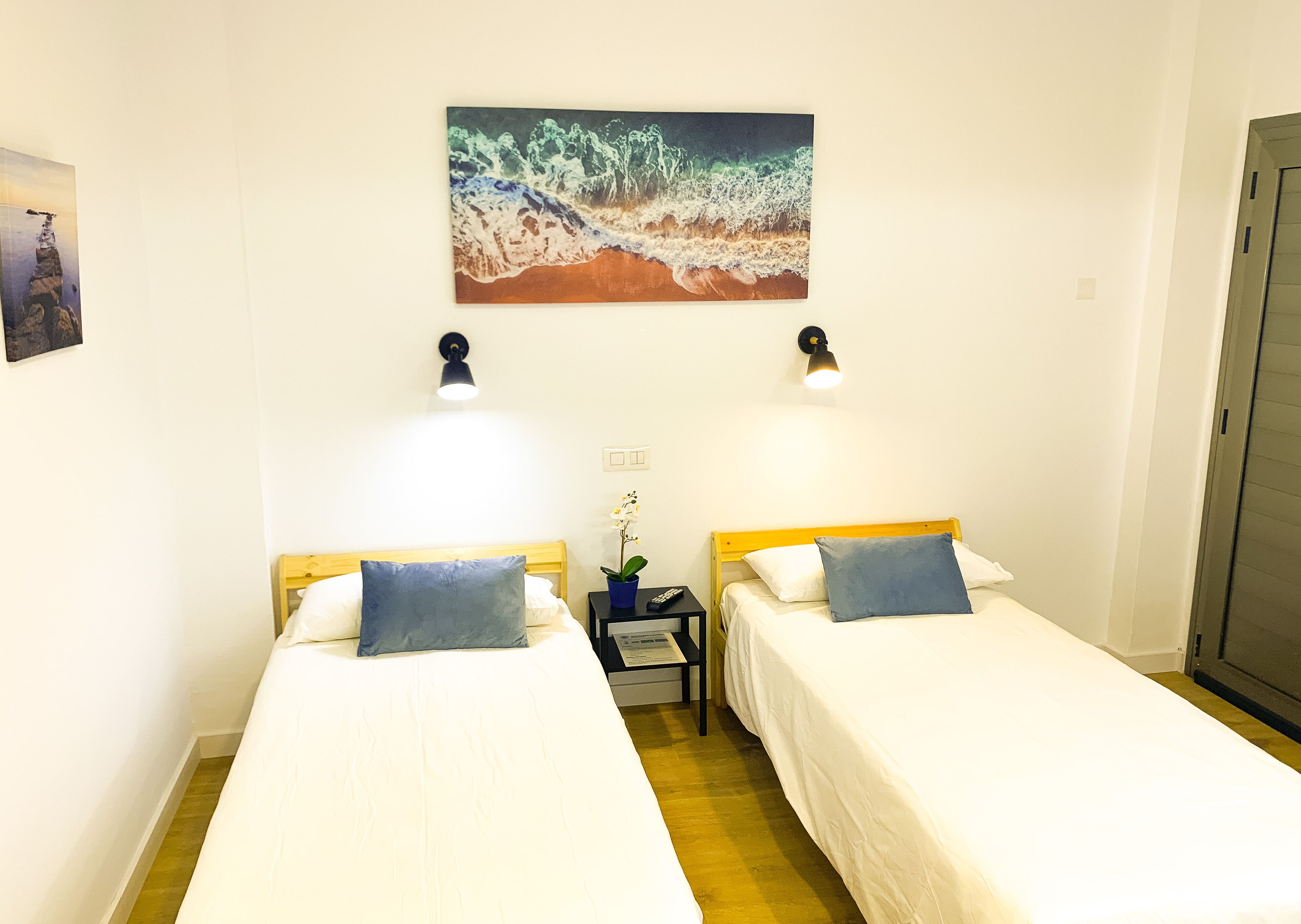Habitación de hotel con dos camas individuales, una mesita de noche con lámpara, un cuadro en la pared y una puerta a la derecha.