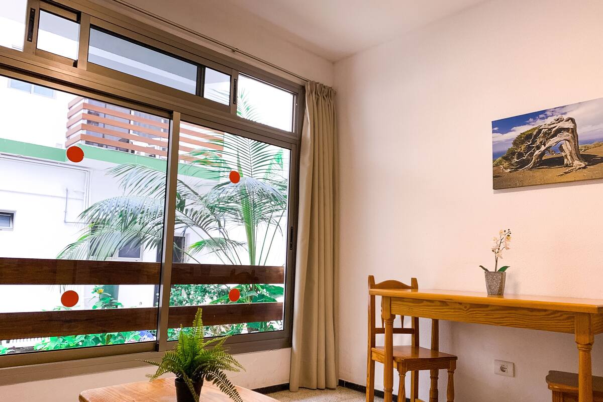 A room with a large window, a wooden table, a chair, a potted plant, and a painting on the wall.