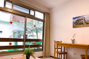 Una habitación con un ventanal grande, una mesa de madera, una silla, una planta en maceta y un cuadro en la pared.