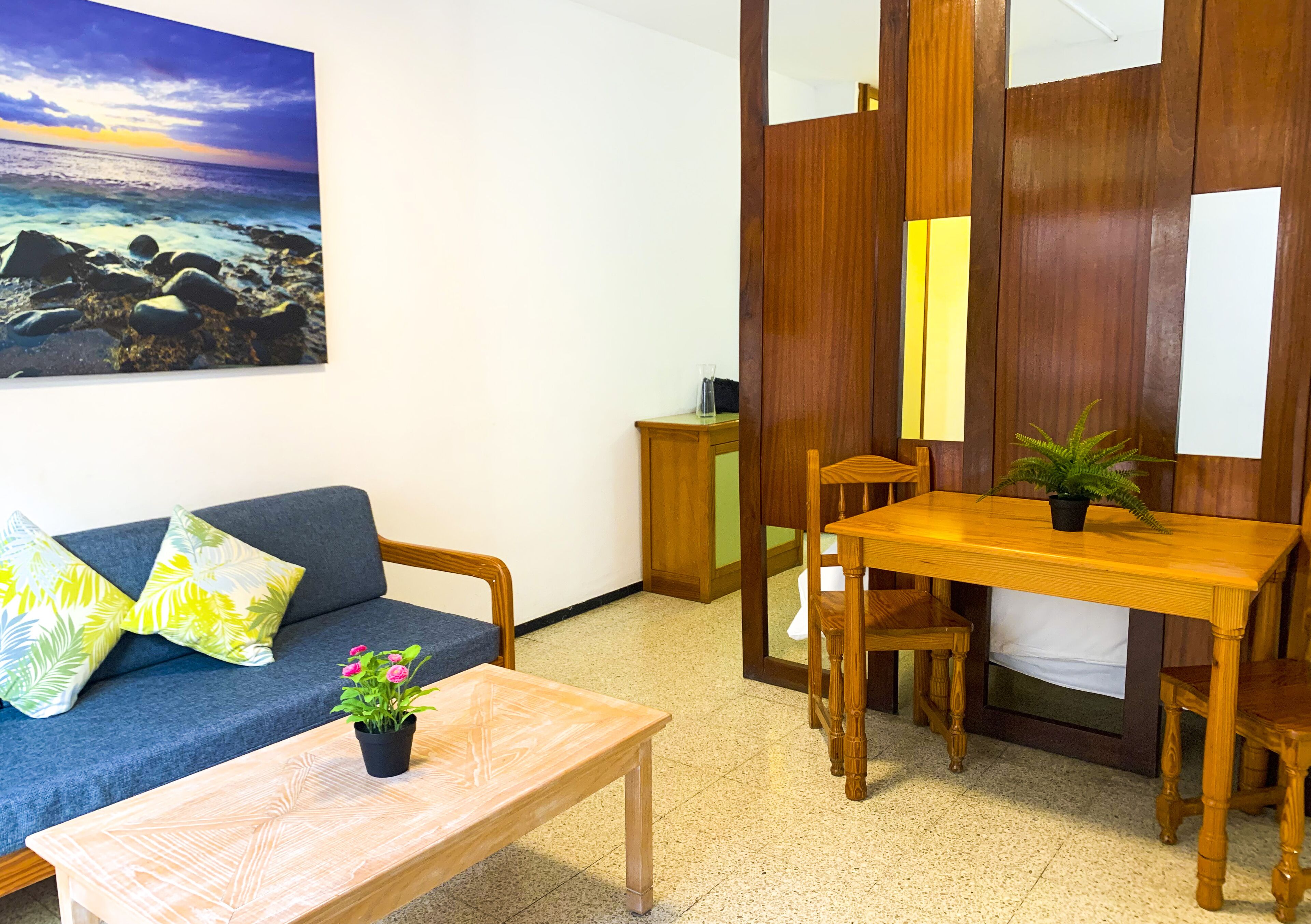 Una habitación con sofá, una mesa de madera y sillas. Hay un cuadro en la pared y una planta sobre la mesa.