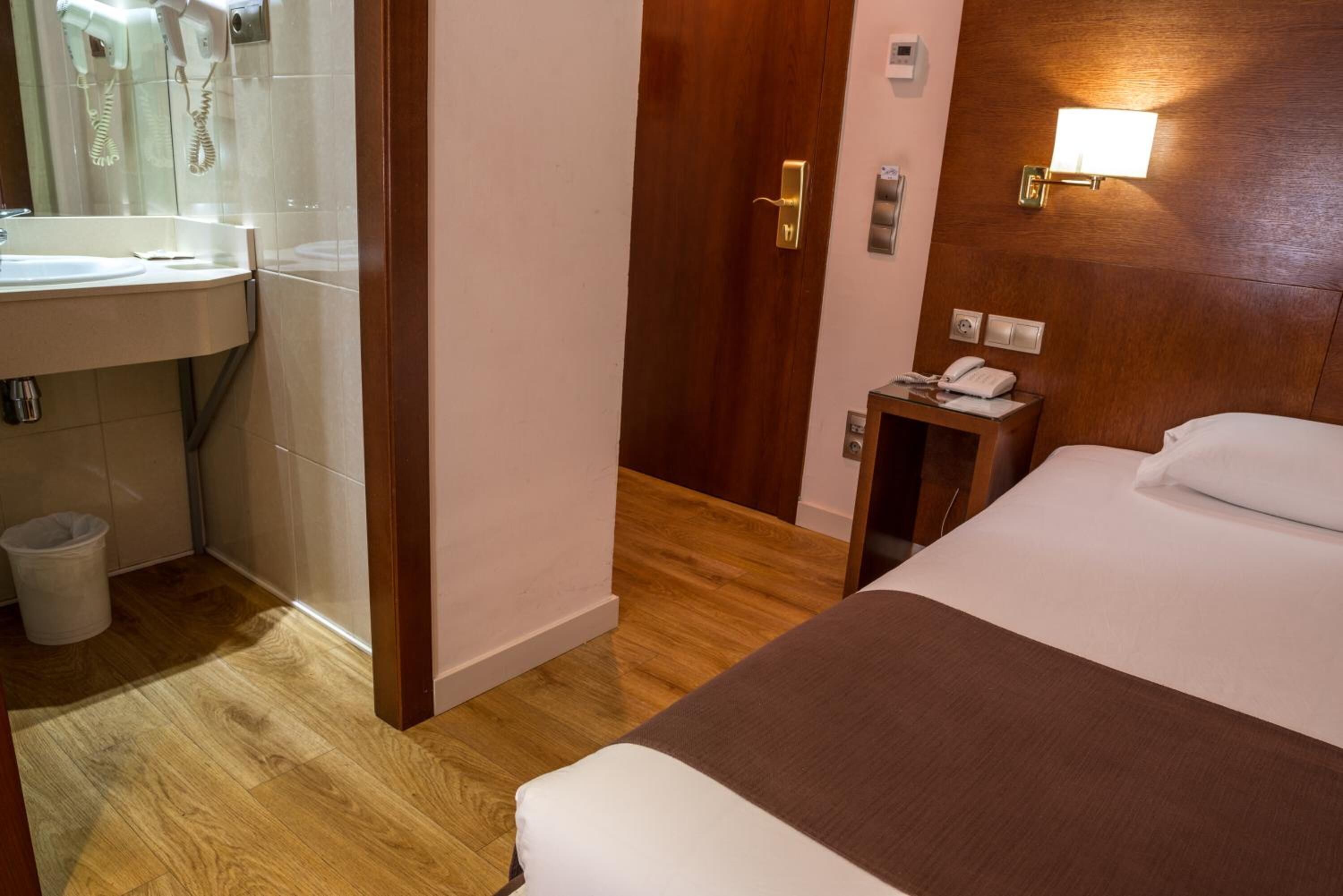 A hotel room with a wooden floor, a bed with a brown bedspread, a bedside table with a phone, and a bathroom with a sink and mirror.