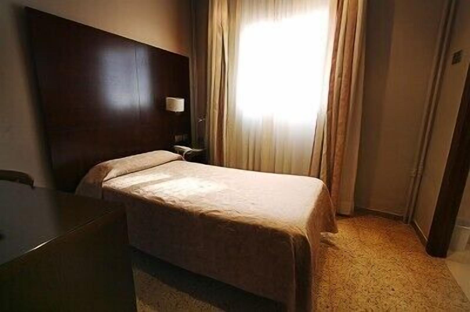 Quarto de hotel com uma cama de solteiro, uma mesa de cabeceira, uma luminária e uma janela com cortinas.
