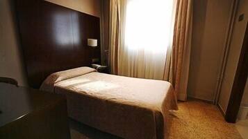 Quarto de hotel com uma cama de solteiro, uma mesa de cabeceira, uma luminária e uma janela com cortinas.
