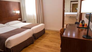 A hotel room with two beds, a wooden desk, a mirror, and a window with curtains.