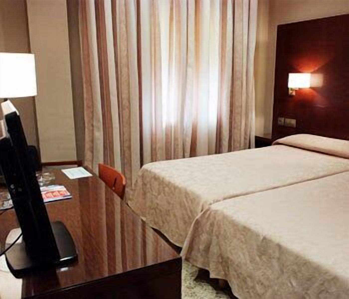 A hotel room with a wooden desk, a lamp, a bed with a beige bedspread, and a curtain with vertical stripes.