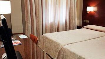 Habitación de hotel con un escritorio de madera, una lámpara, una cama con colcha color beige y una cortina a rayas verticales.